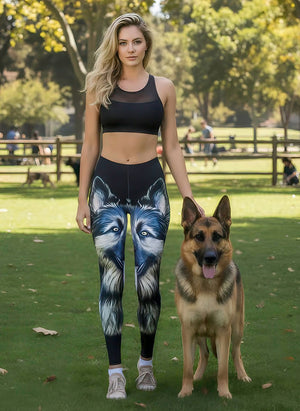 Woman in a black sports bra wearing German Shepherd leggings with a large blue-eyed shepherd face, beside a real German Shepherd at the park.