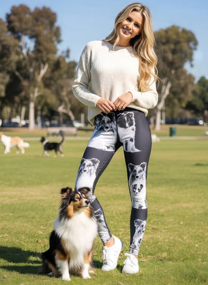 Woman in a cream knit sweater wearing Shetland print leggings with Shetland Sheepdog faces on black, standing next to a real Sheltie at the park.