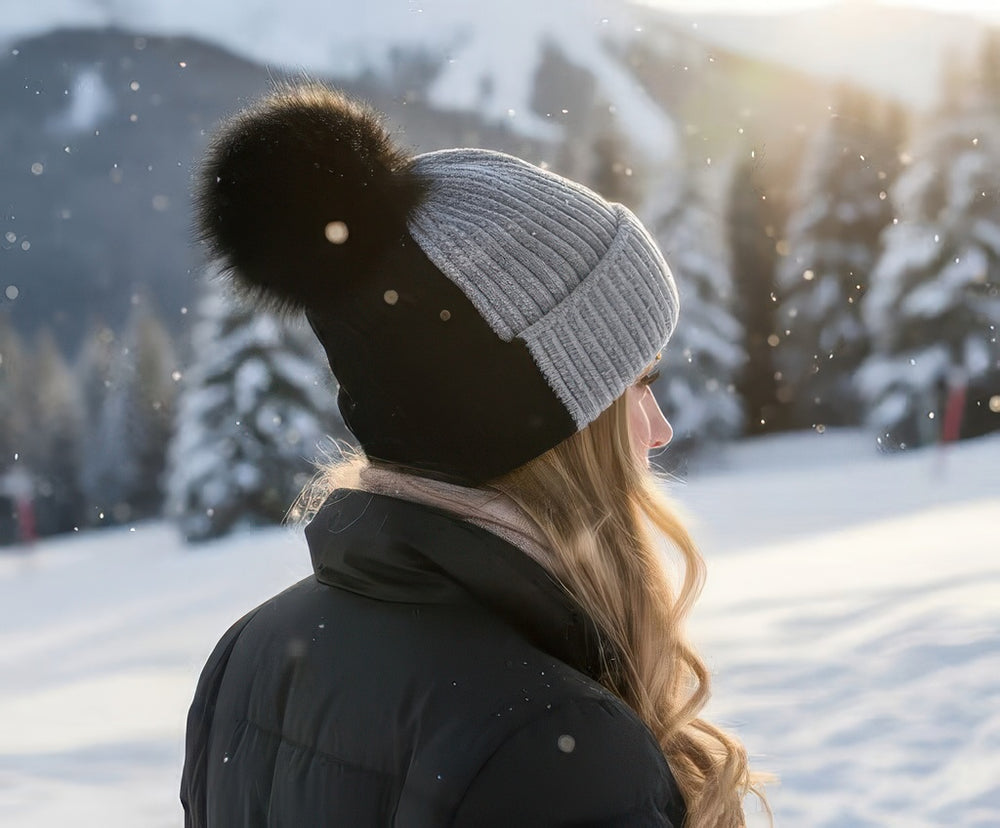 Side view of a woman wearing the gray and black Vail knit beanie with black fox pom, snowy forest backdrop.