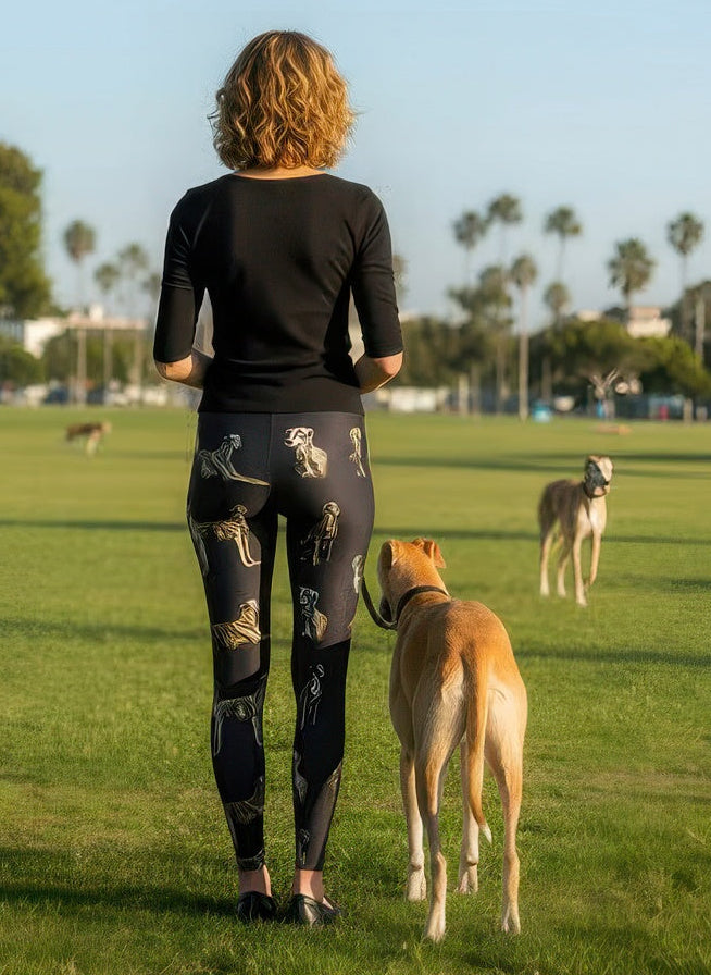 Back view of woman wearing Denimaxx Whippet print leggings with a tan whippet on a leash beside her at a dog park with palm trees.