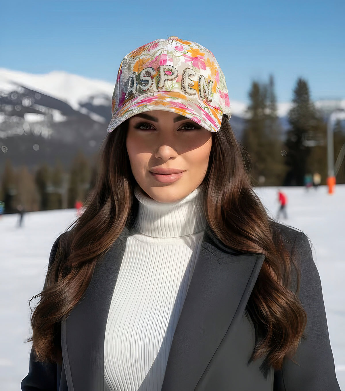 Woman wearing the Pearl-Detail Aspen baseball cap with pearl ASPEN lettering on floral lace, standing on a snowy ski slope.
