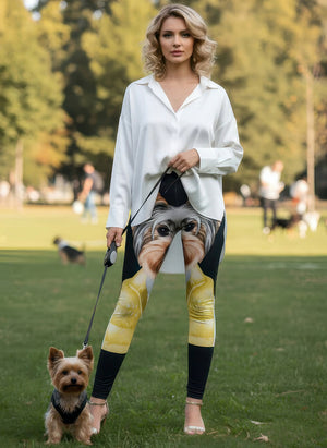 Woman in a white shirt wearing Yorkie print leggings with a large Yorkie face artwork, holding a leash with a real Yorkie in a grassy park.