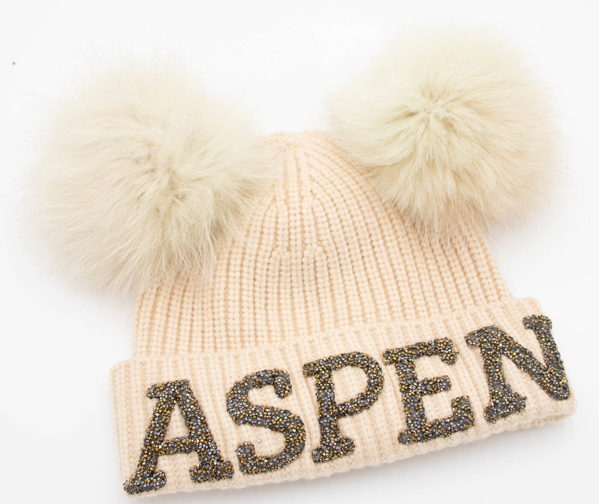 Cream knit Aspen beanie with dark gold crystal ASPEN letters and two cream fox fur poms, flat product shot on white.