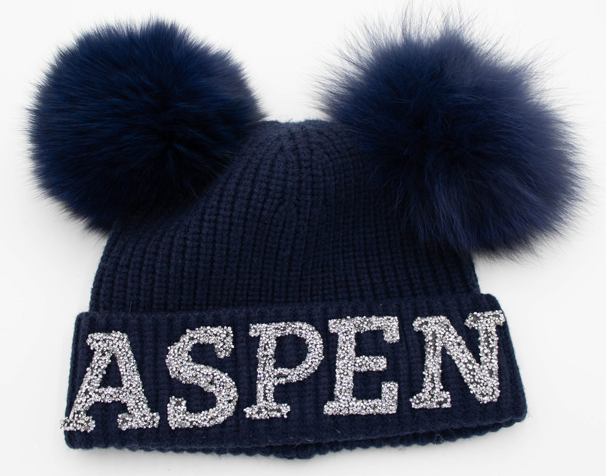Navy knit Aspen ski beanie with sparkling ASPEN letters and two navy fox fur poms, flat product shot on white.