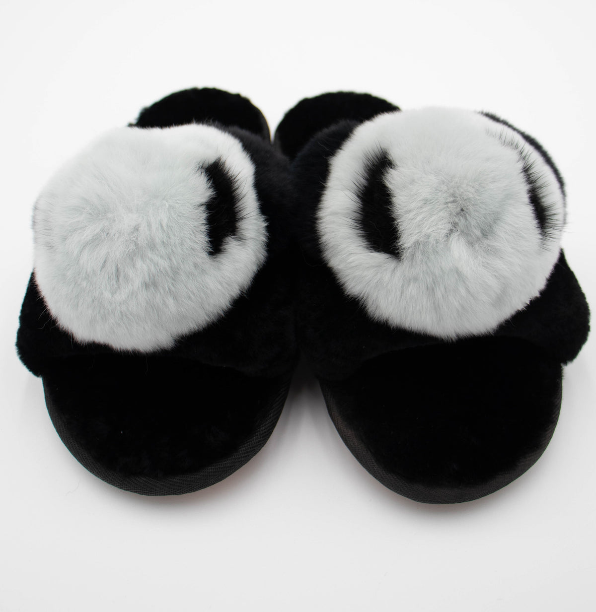 Pair of black sheepskin slide slippers with fluffy white and gray fur pom tops, front view.