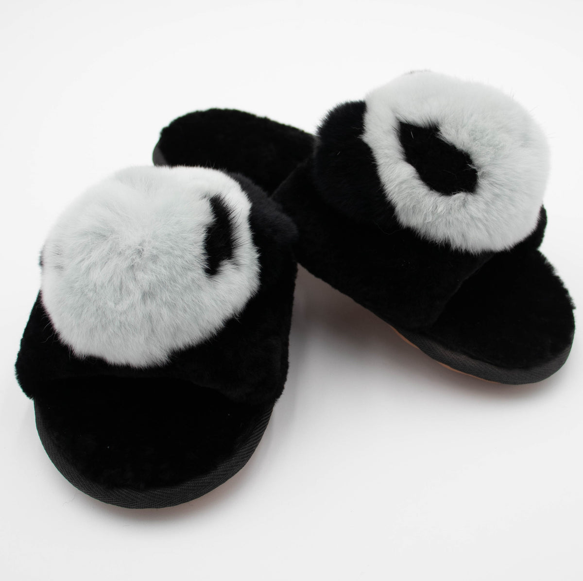 Angled view of black sheepskin slide slippers with white fur pom tops and plush black footbeds.