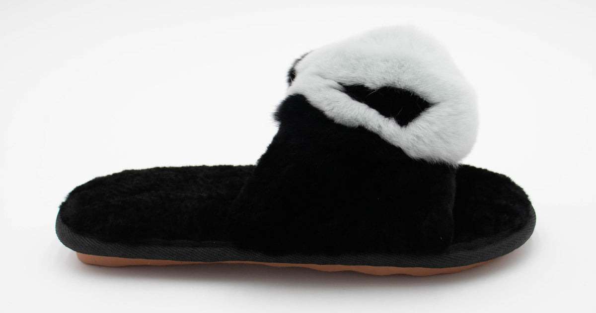 Side profile of a single black sheepskin slide slipper with white fur pom top and plush shearling footbed.