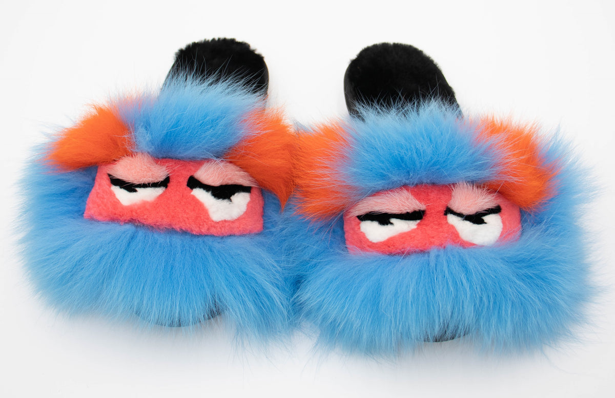 Top view of blue sheepskin slide slippers with pink fox monster-face trim and orange accents, studio shot.