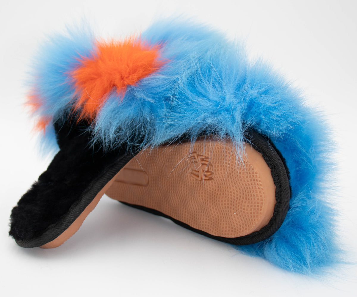 Angled view showing the tan rubber sole of the blue sheepskin slippers with blue fox trim and orange monster-face accents.