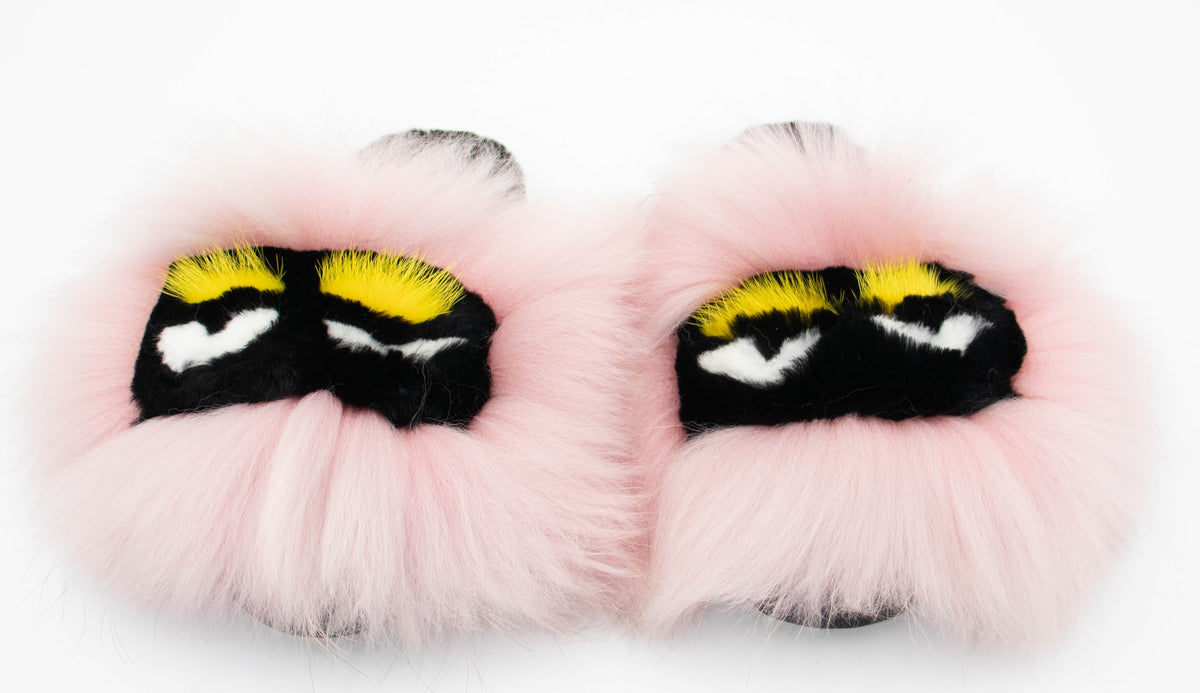 Pair of pink fox fur slide slippers with monster-eye embroidery in yellow, black, and white, front view.