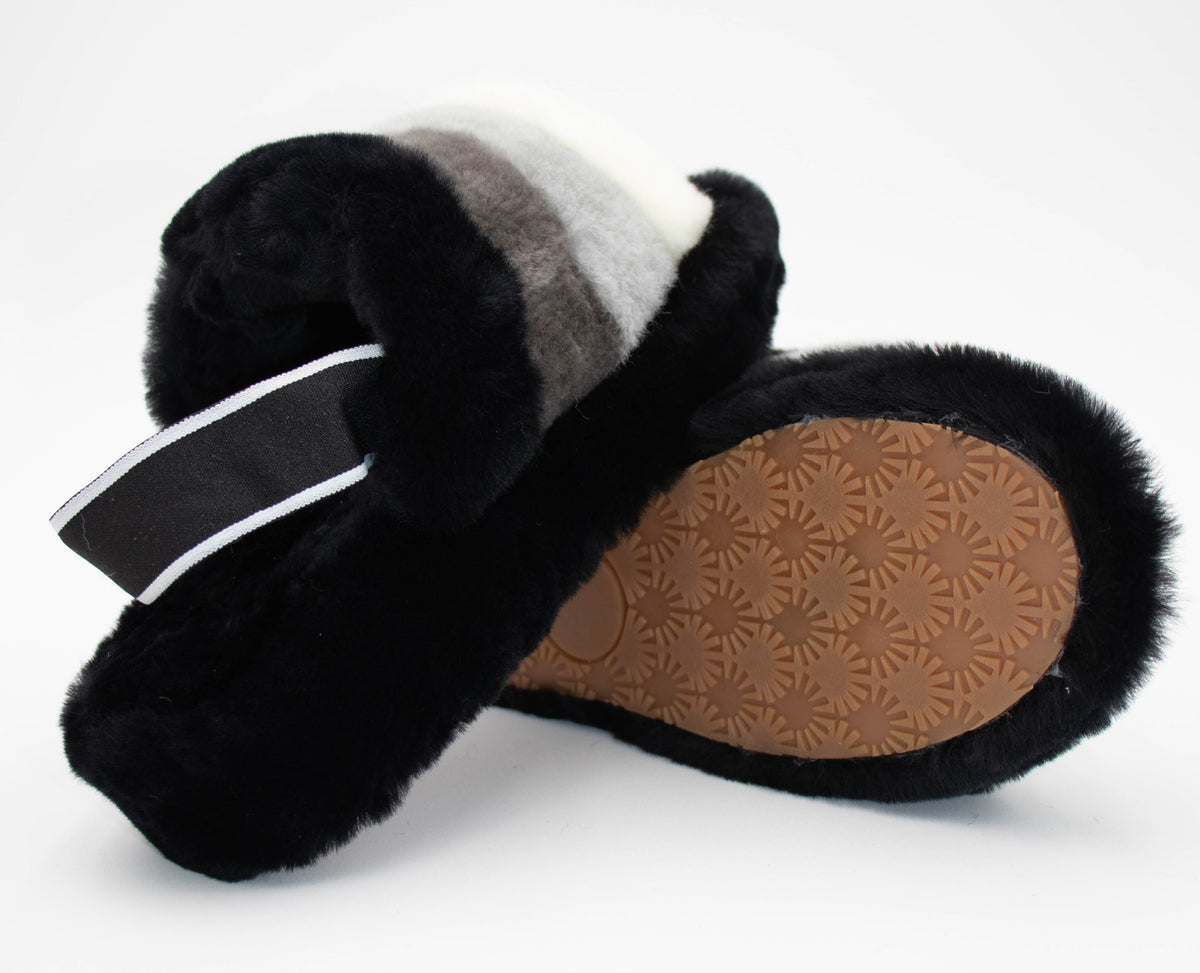 Angled view showing the brown patterned rubber sole of the black sheepskin slide slippers with gray and white stripes.