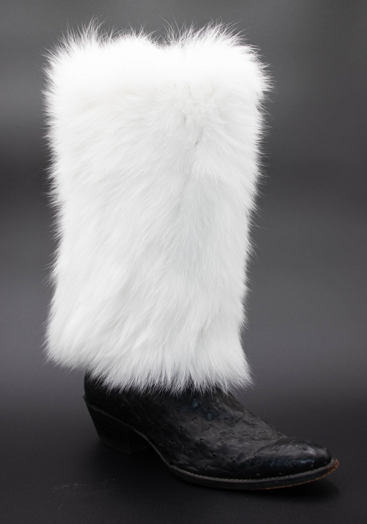 White fox fur leg warmer shown on a black leather boot, fluffy white fur, dark studio background.