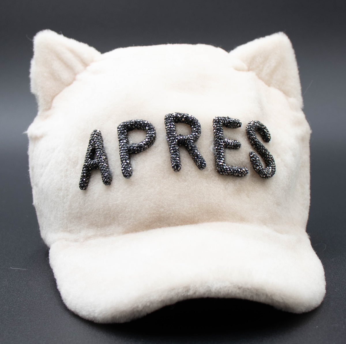 Front view of the white Apres Crystal cat ear hat with dark crystal APRES lettering on a dark background.