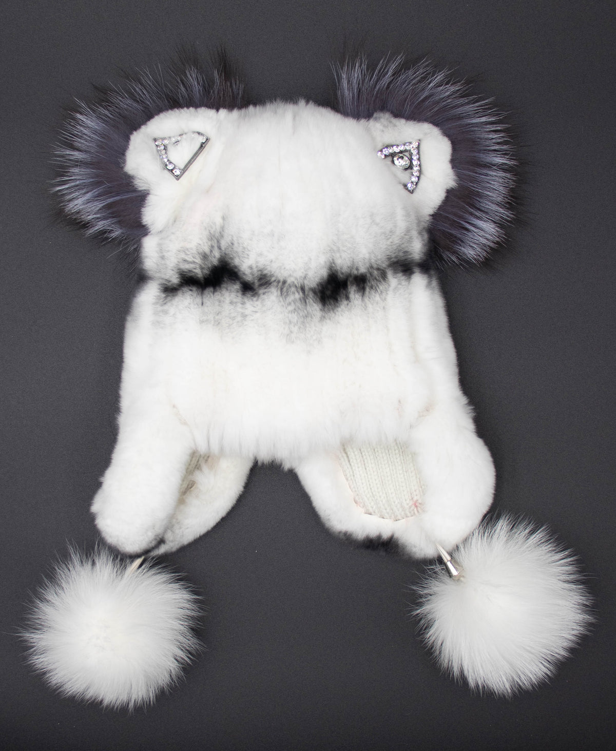 Front view of the kids white rex rabbit fur trapper hat with dark fur ear tufts, rhinestone details, and white pom-poms, on black.