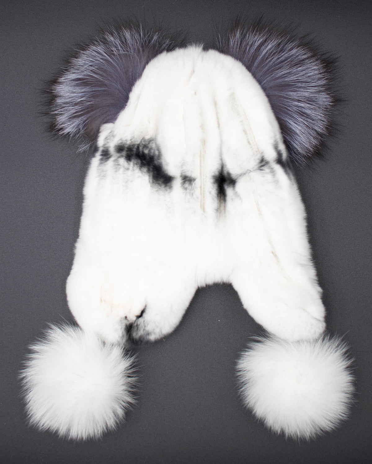 Back view of the kids white rex rabbit fur trapper hat with dark ear tufts and white pom-pom ties, studio shot on black.