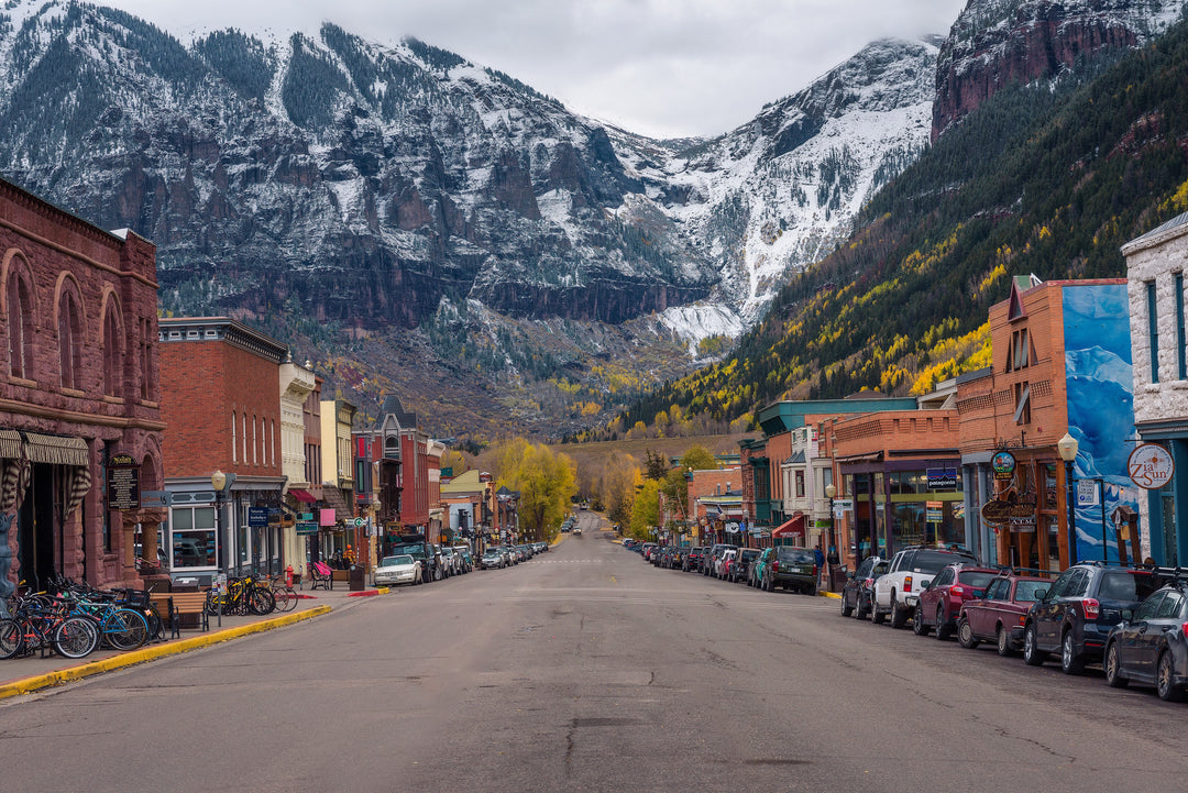 Denimaxx MAG 628 Telluride photo print of the historic main street with brick storefronts, parked cars, and snow-dusted peaks behind.