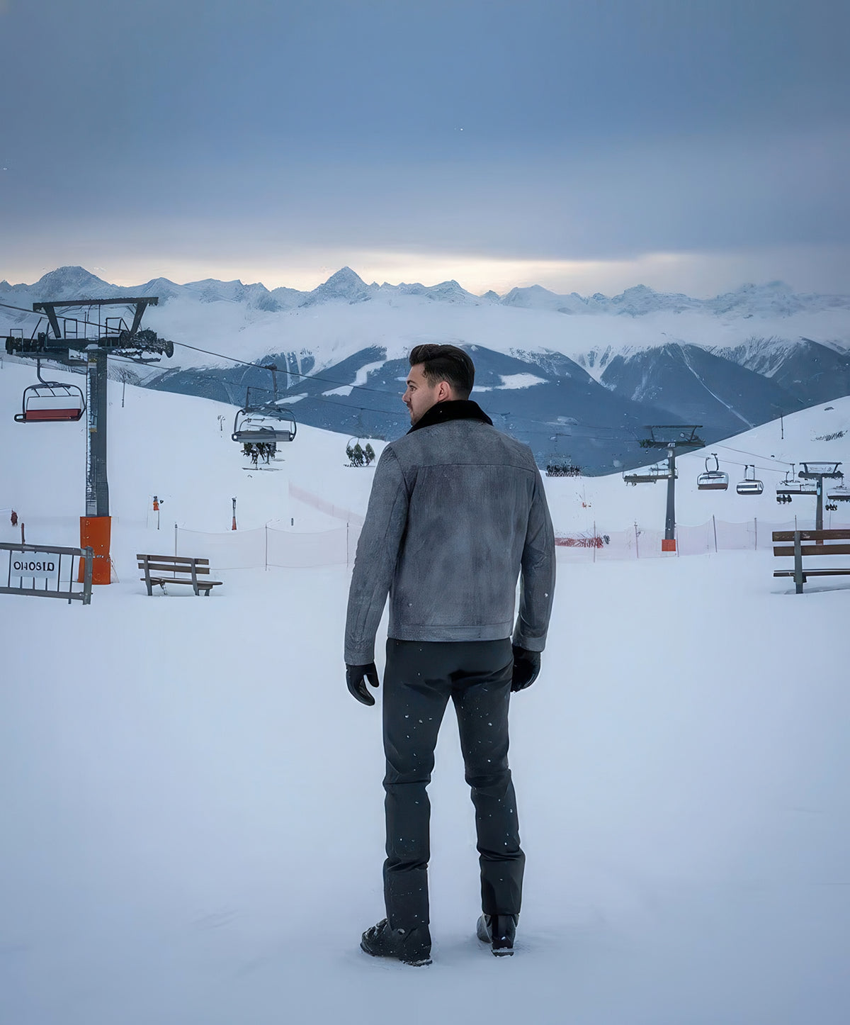 Back view of man wearing the gray leather shearling lined jacket on a snowy ski slope with chairlifts and mountain peaks behind.