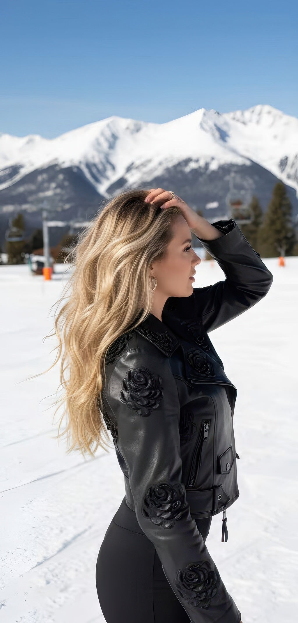 Side view of woman in the black leather jacket with embroidered black roses on the shoulders and sleeves, snowy mountain backdrop.