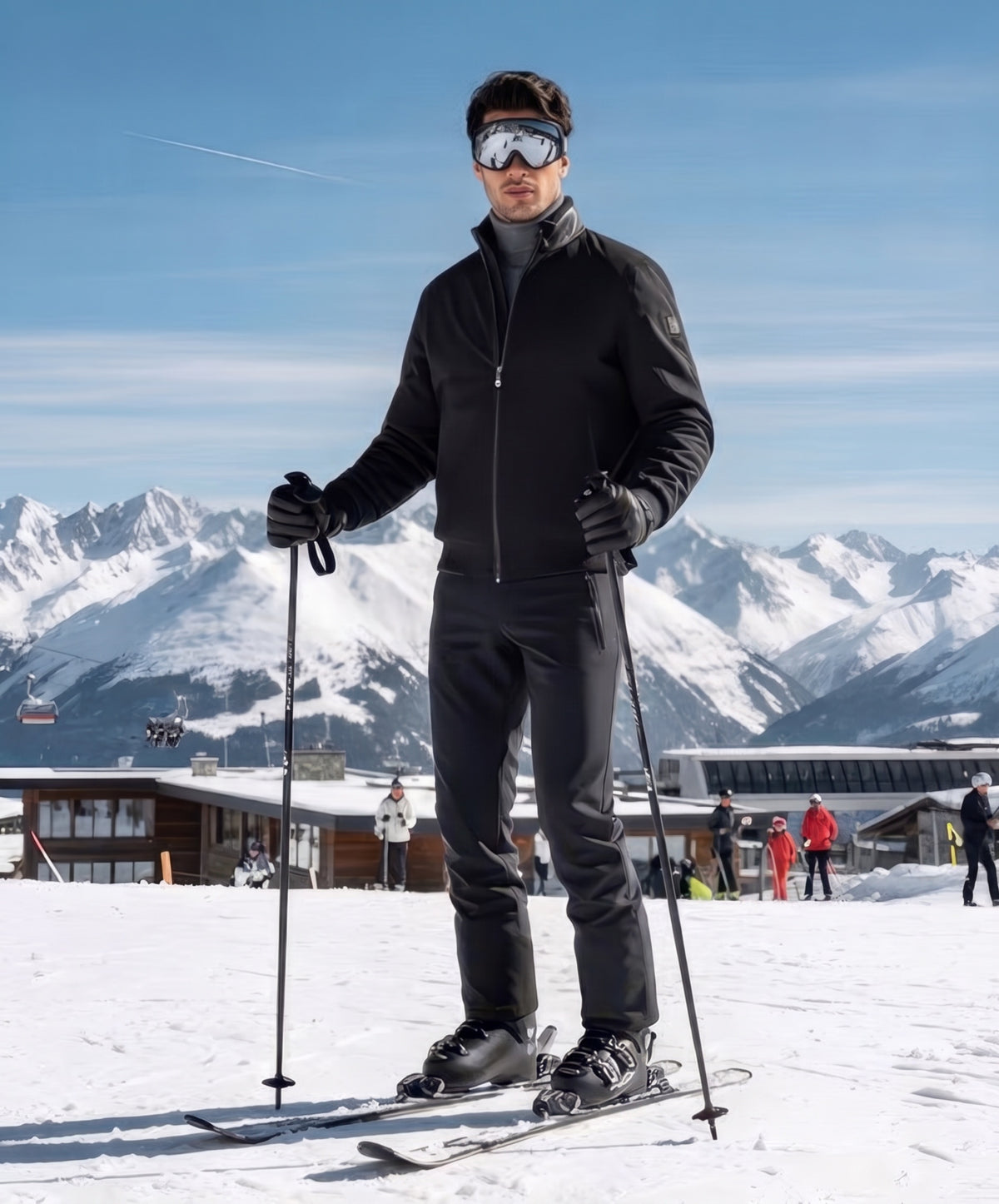 Man on skis holding poles, wearing the black nylon jacket with shearling and ski goggles, snowy mountain resort behind.