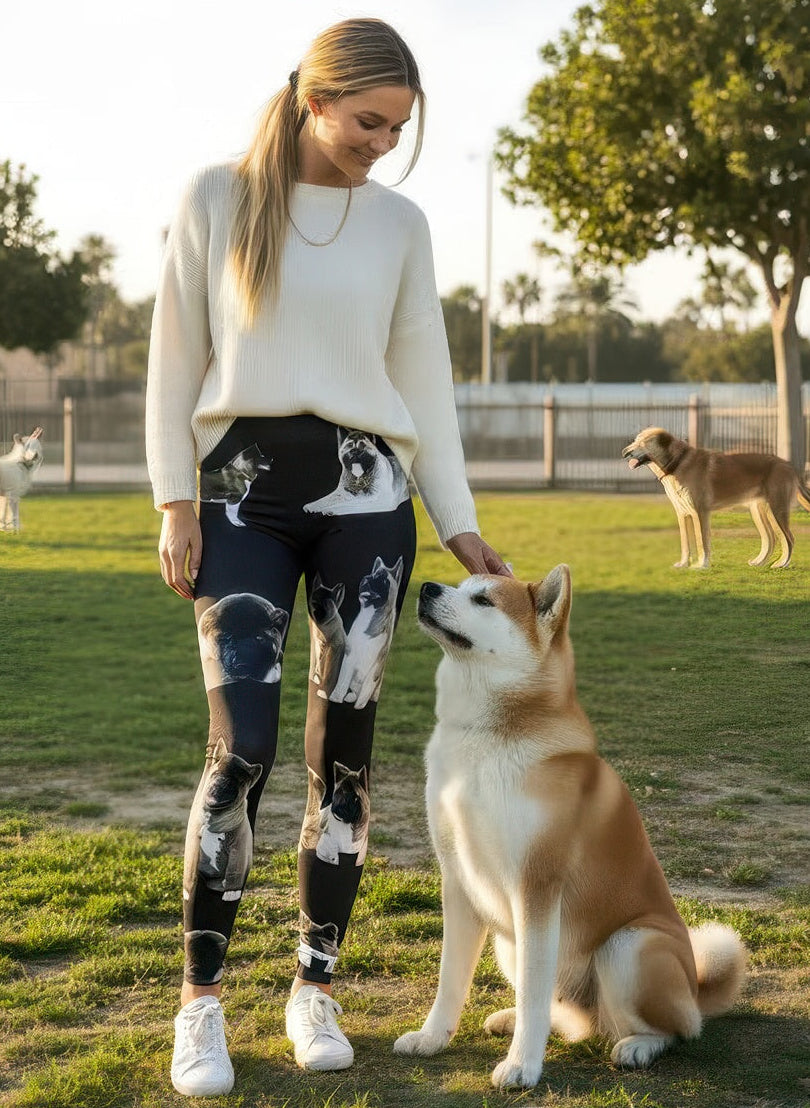 Woman smiling and petting a seated Akita at a dog park while wearing Denimaxx Akita print high-rise leggings with realistic Akita faces.