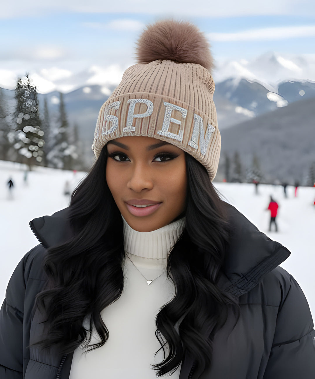 Woman wearing the tan Aspen knit beanie with silver crystal ASPEN lettering and a tan fox pom on a busy ski slope with skiers.