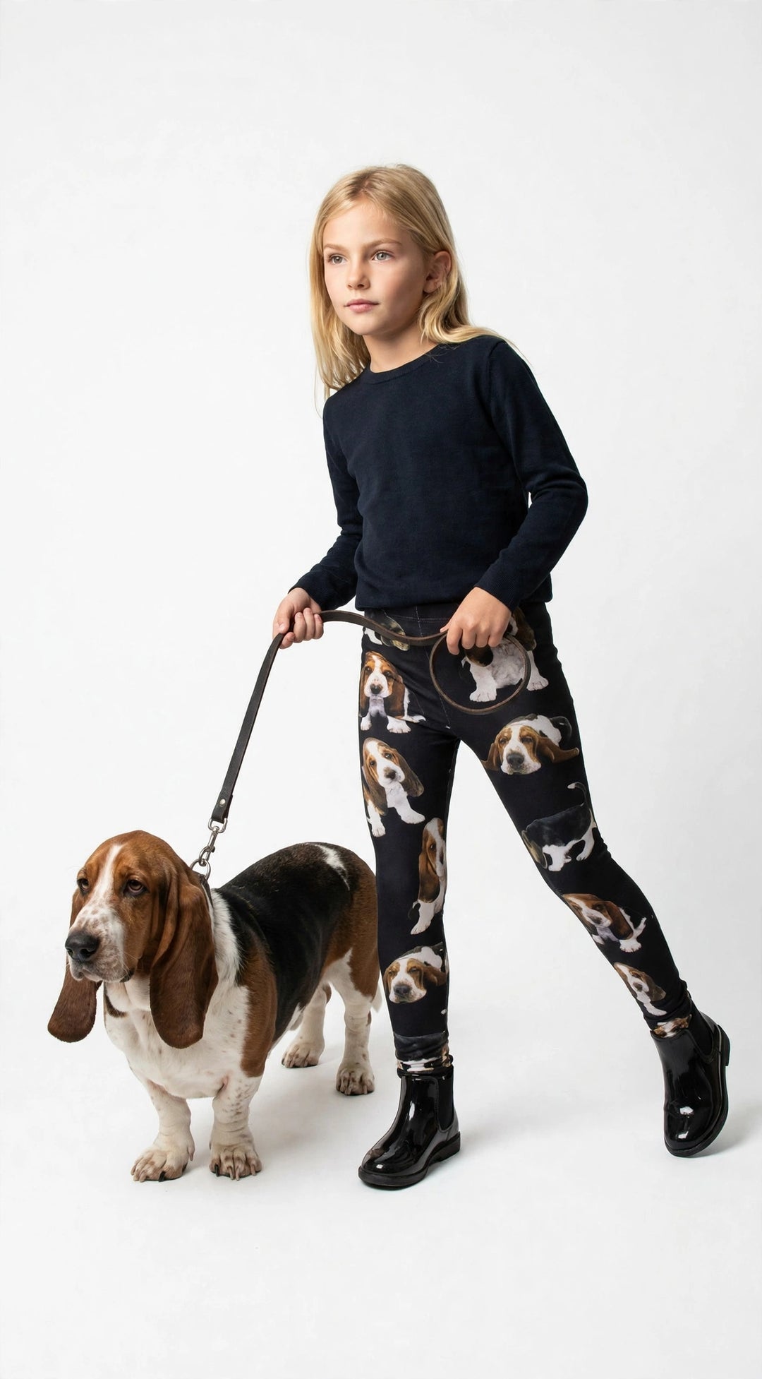 Young girl walking a real Basset Hound on a leash, wearing Denimaxx kids Basset Hound print leggings with puppy prints, black boots, white studio.