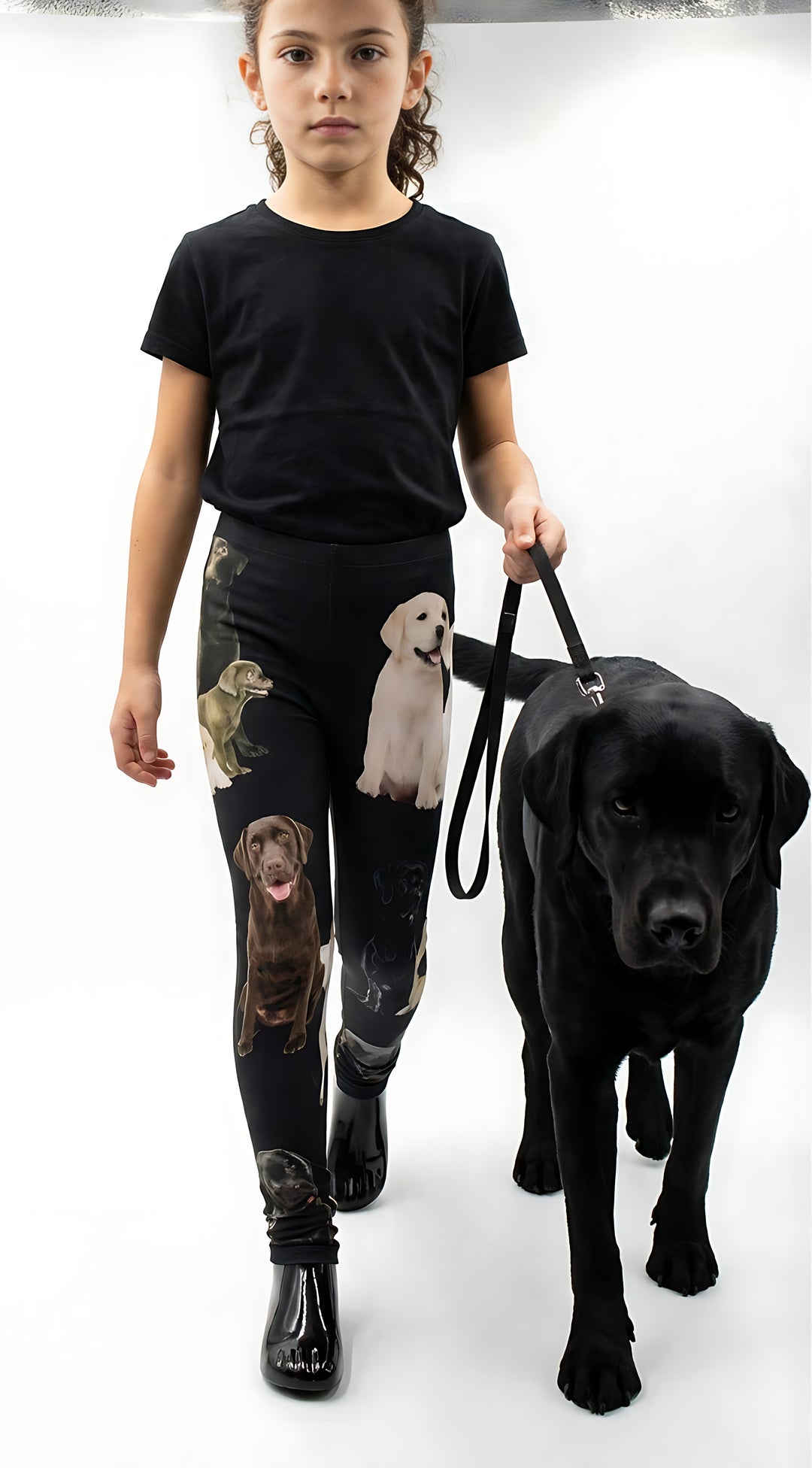 Young girl walking a real black Labrador on a leash while wearing kids Labrador print leggings and a black t-shirt in a white studio.