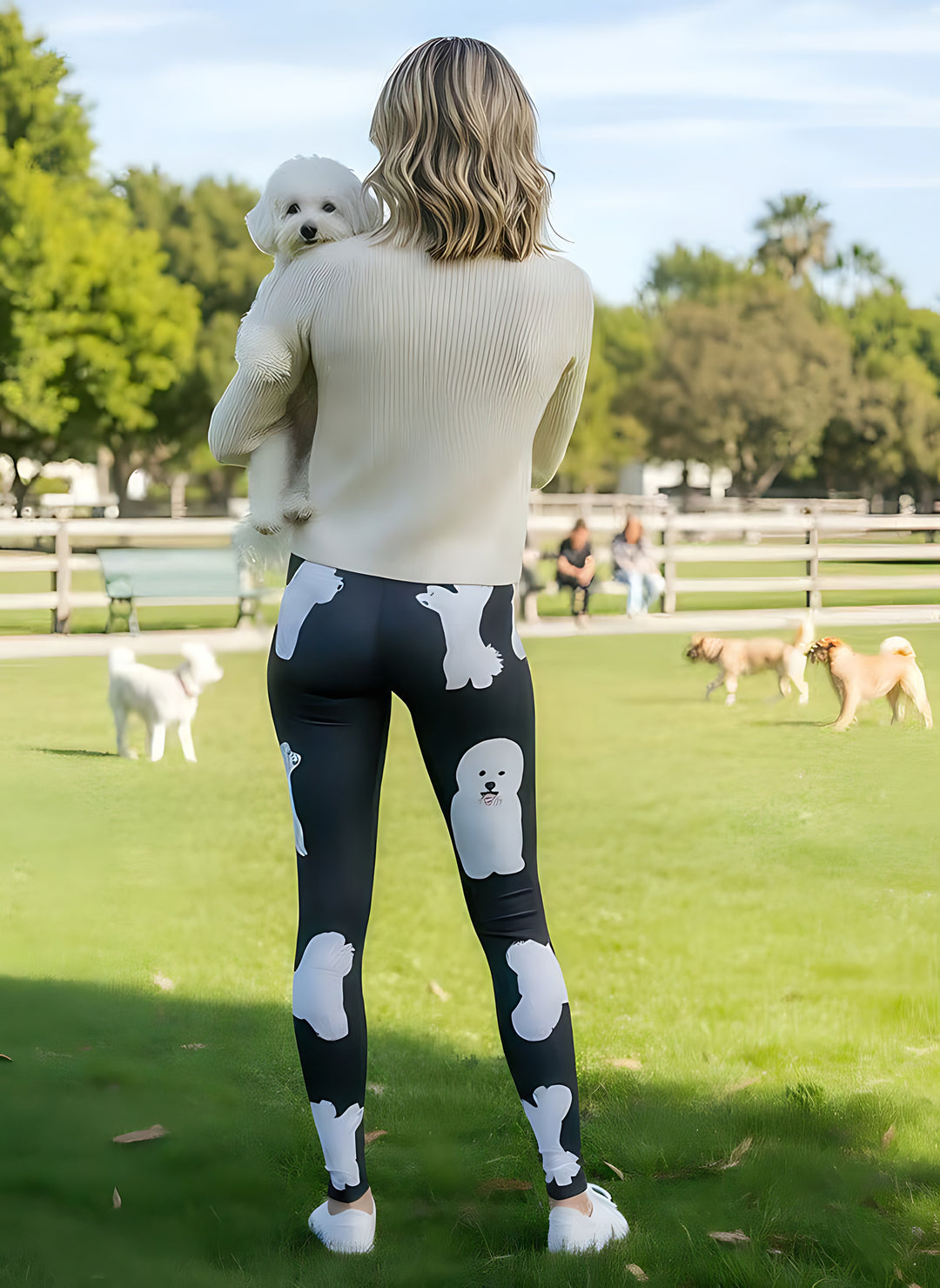 Back view of woman holding a Bichon Frise over her shoulder, wearing Bichon Frise print leggings at a sunny dog park.