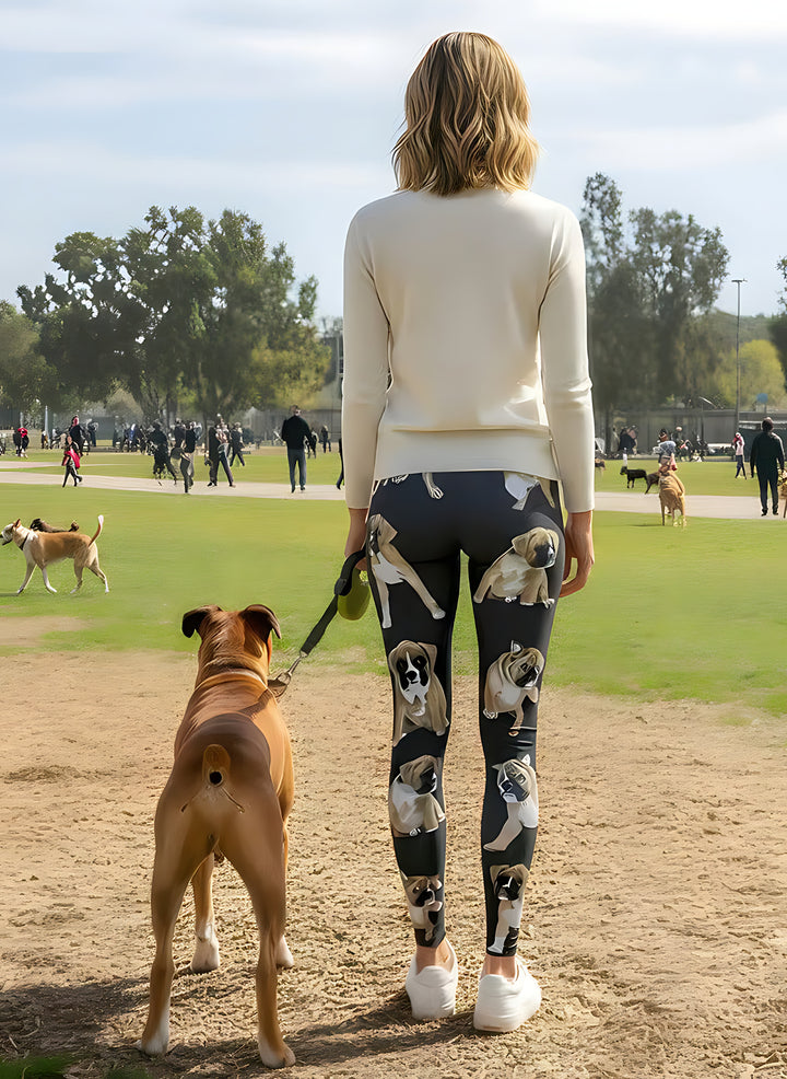 Back view of woman in a cream sweater wearing Boxer print leggings, holding a leash with a real Boxer dog at a dog park.