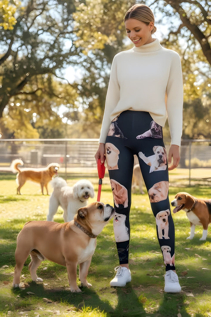 Woman wearing Mixed Dog Breed print leggings with assorted dog faces, cream turtleneck, holding a leash as a bulldog sniffs her leg at a dog park.