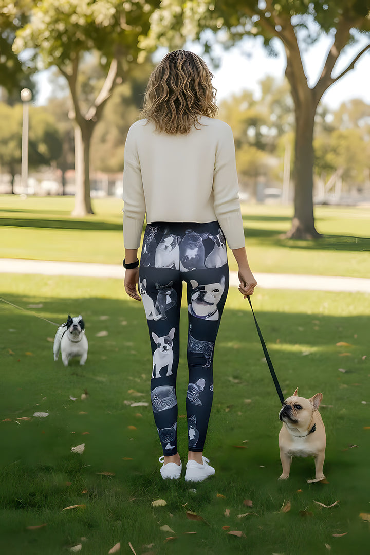 Back view of woman in a cream sweater wearing French Bulldog print leggings, walking real French bulldogs on leashes in a park.