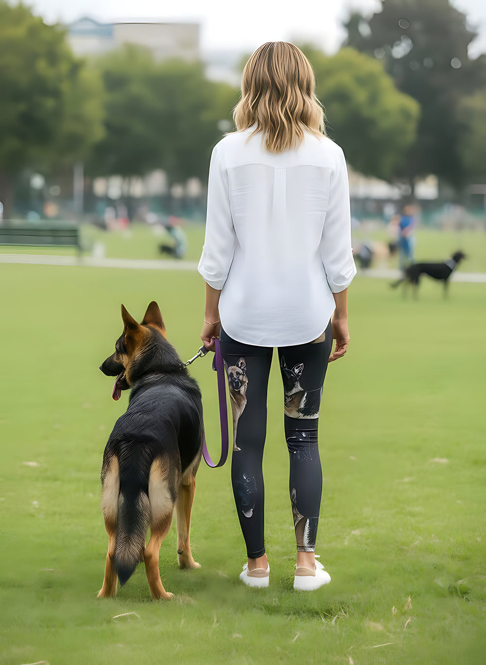 Back view of woman in a white shirt wearing German Shepherd print leggings, holding a real German Shepherd on a purple leash in a green park.
