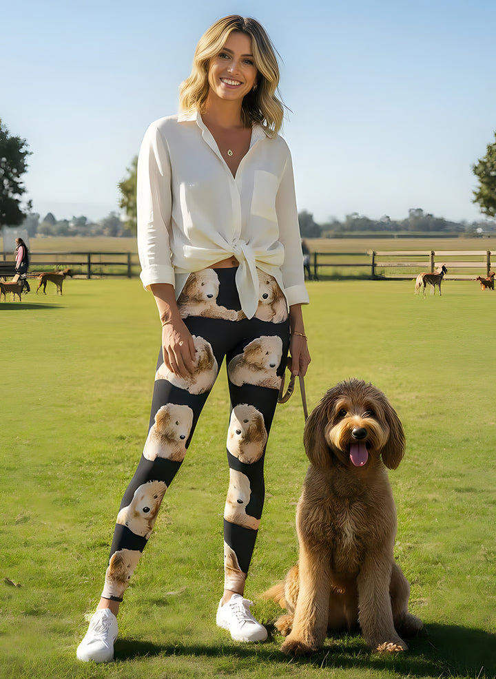 Woman in a tied white shirt wearing Goldendoodle print leggings with fluffy doodle faces, holding a real Goldendoodle on a leash in a sunny field.