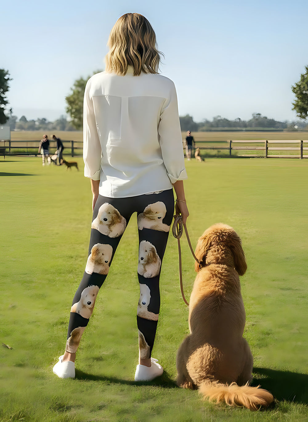 Back view of woman wearing Goldendoodle print leggings, standing with a real Goldendoodle on a leash in a green field.