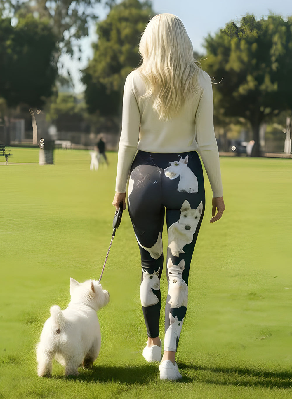 Back view of woman at a dog park wearing Scottish Terrier print leggings with white terrier faces on black, walking a real white terrier on a leash.