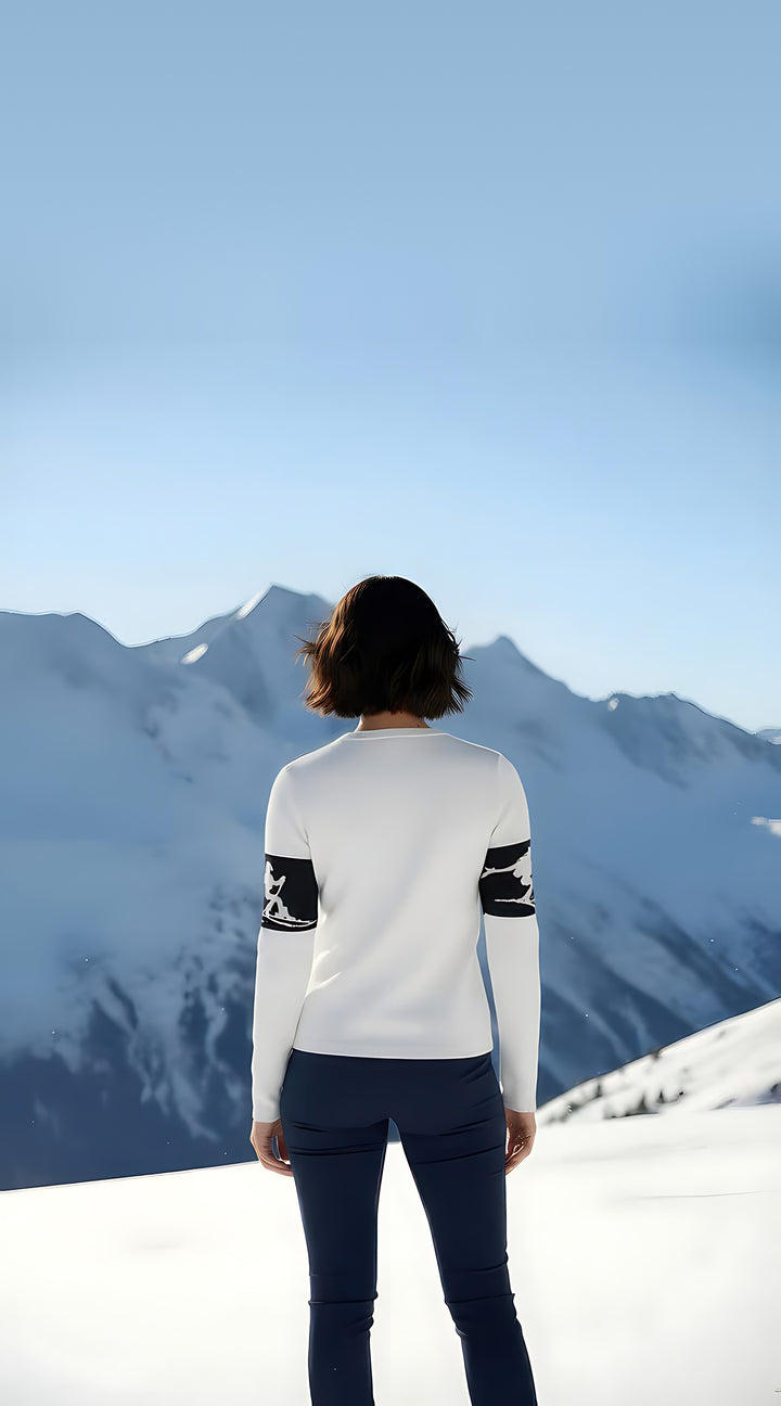 Back view of woman wearing the white Cowgirl Skiers intarsia crewneck sweater with skier sleeve bands, facing snowy peaks.