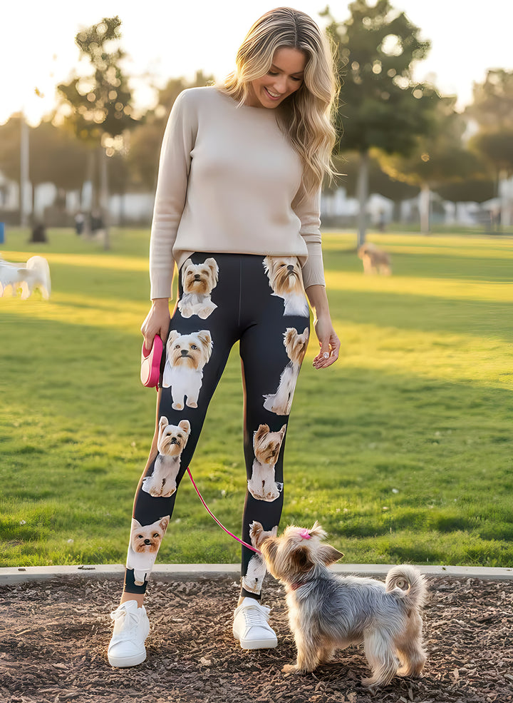 Smiling woman in a beige sweater wearing Yorkshire Terrier print leggings with small Yorkie artwork, holding a leash with a real Yorkie at the park.