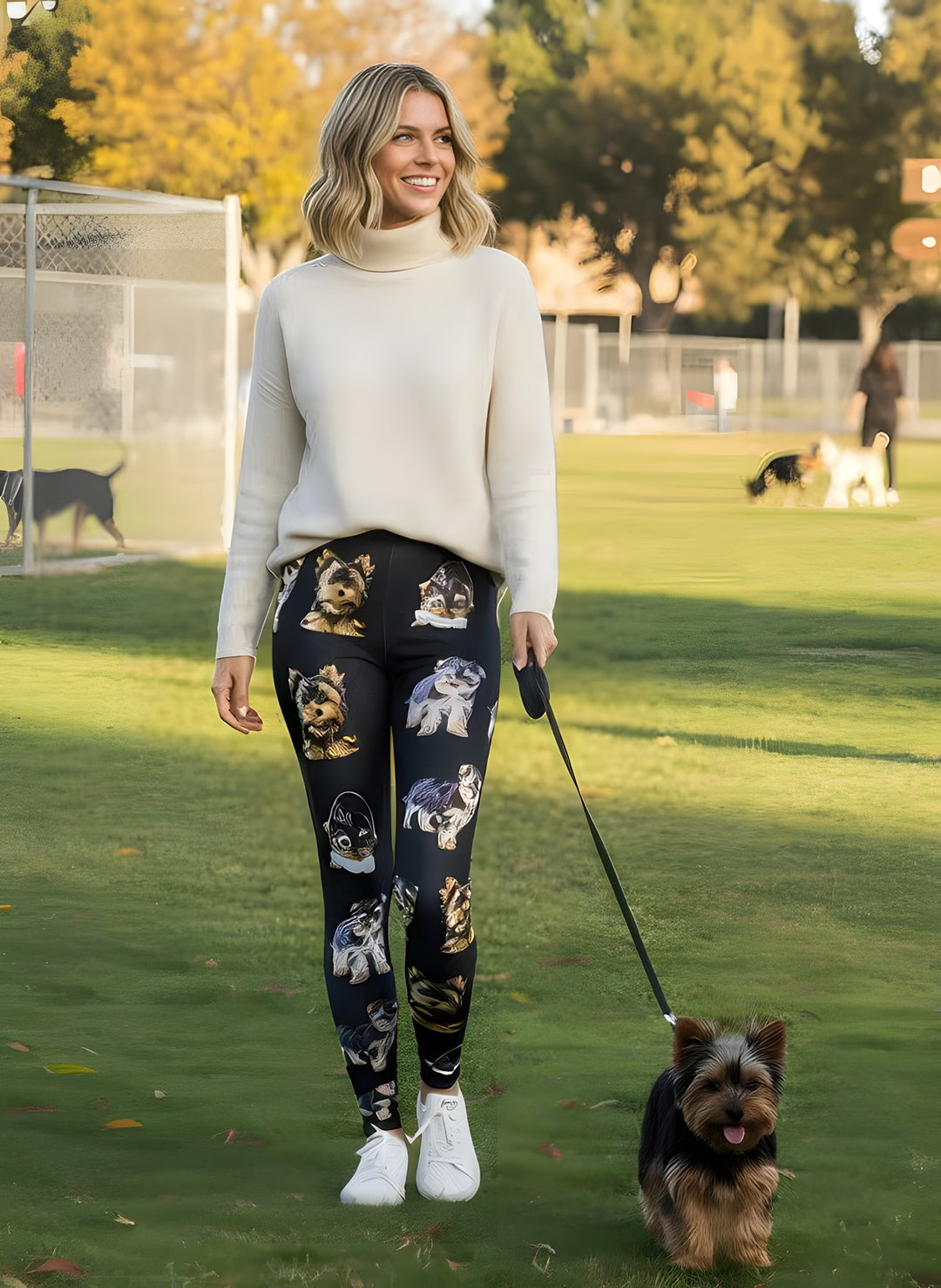 Woman in a cream turtleneck and white sneakers wearing Yorkie print leggings with puppy face artwork, walking a real Yorkie on a leash at the dog park.