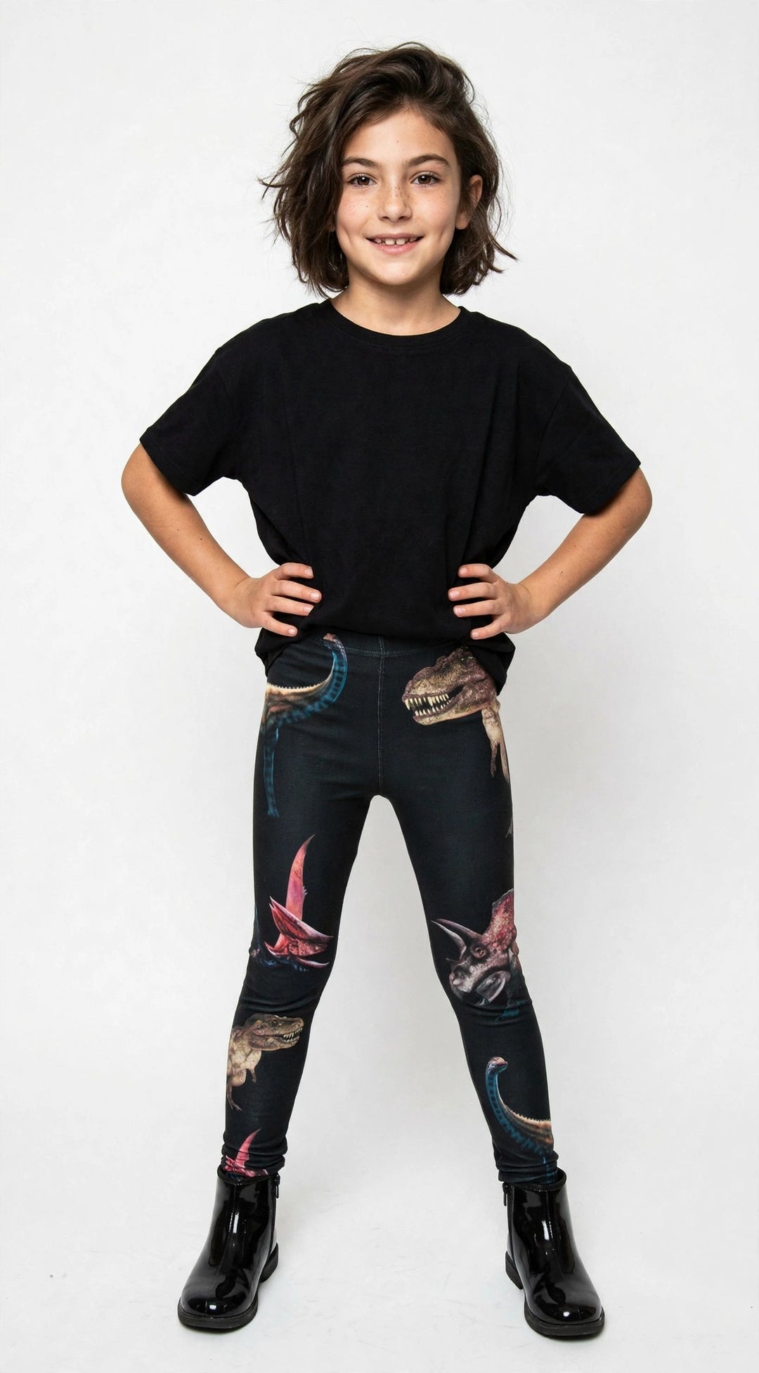 Young girl with hands on hips wearing Denimaxx kids dinosaur print leggings with T-Rex and pterodactyl artwork, black t-shirt and boots on a white background.