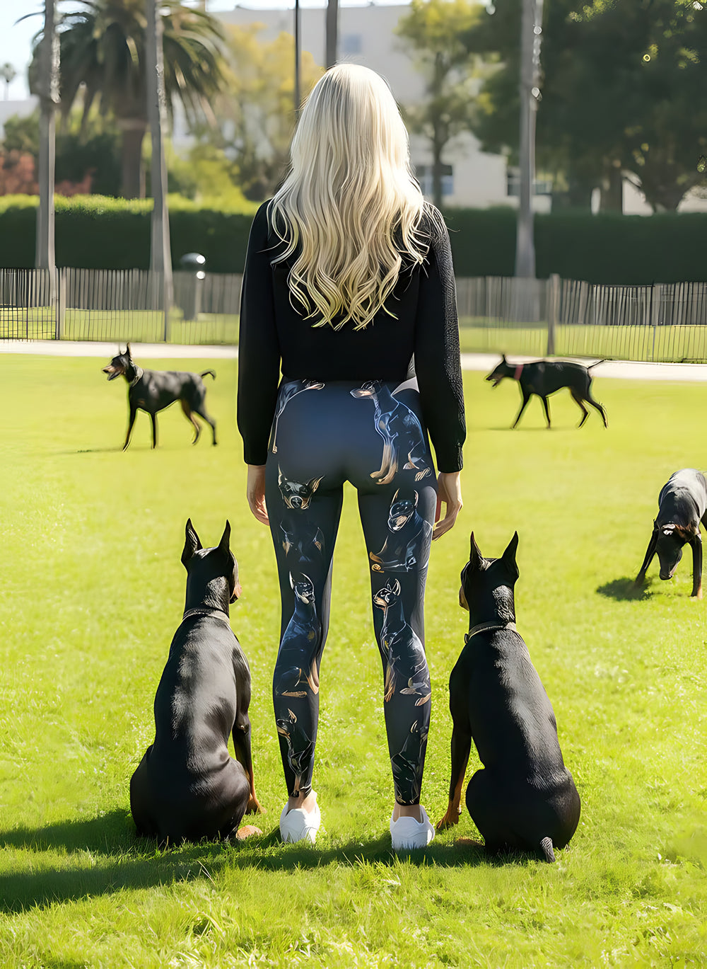 Back view of woman wearing Doberman print leggings with Doberman artwork, standing between two real sitting Dobermans in a sunny park.