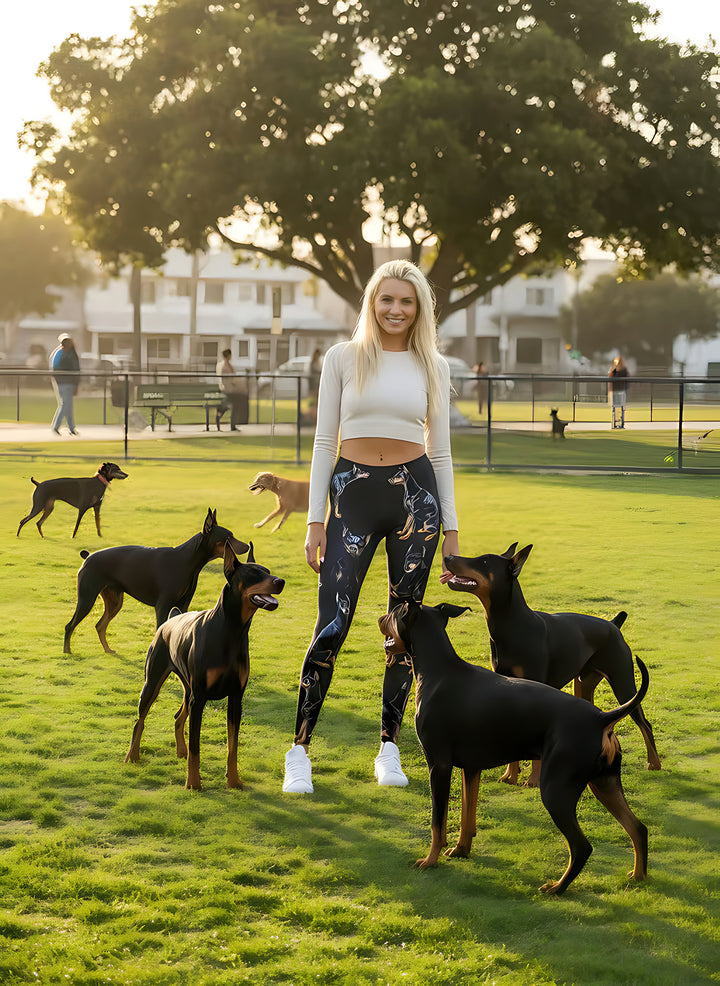 Woman in a white top wearing Doberman print leggings, surrounded by playful real Dobermans at a dog park at sunset.