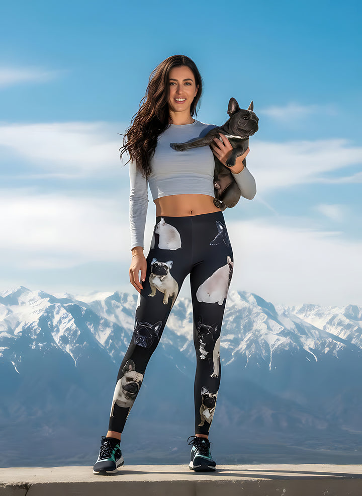 Woman holding a real French Bulldog while wearing French Bulldog print leggings and a grey crop top, snowy mountains behind.