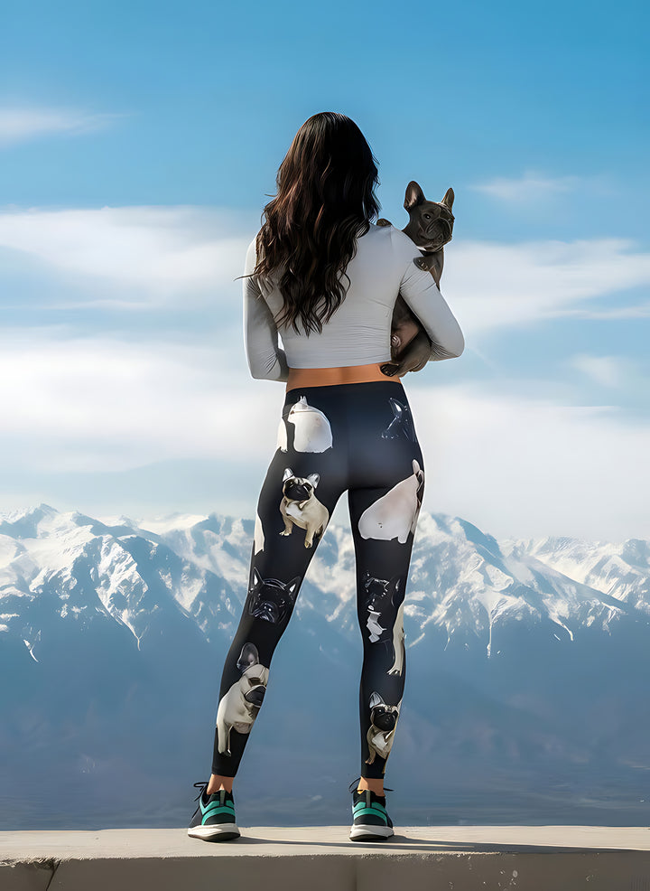 Back view of woman holding a French Bulldog over her shoulder, wearing French Bulldog print leggings, mountain peaks behind.