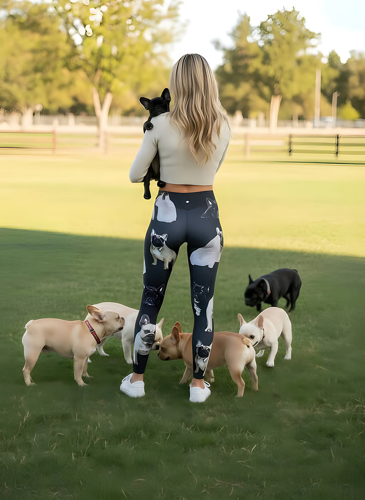 Back view of woman carrying a French Bulldog puppy, wearing French Bulldog print leggings among real Frenchies at a dog park.