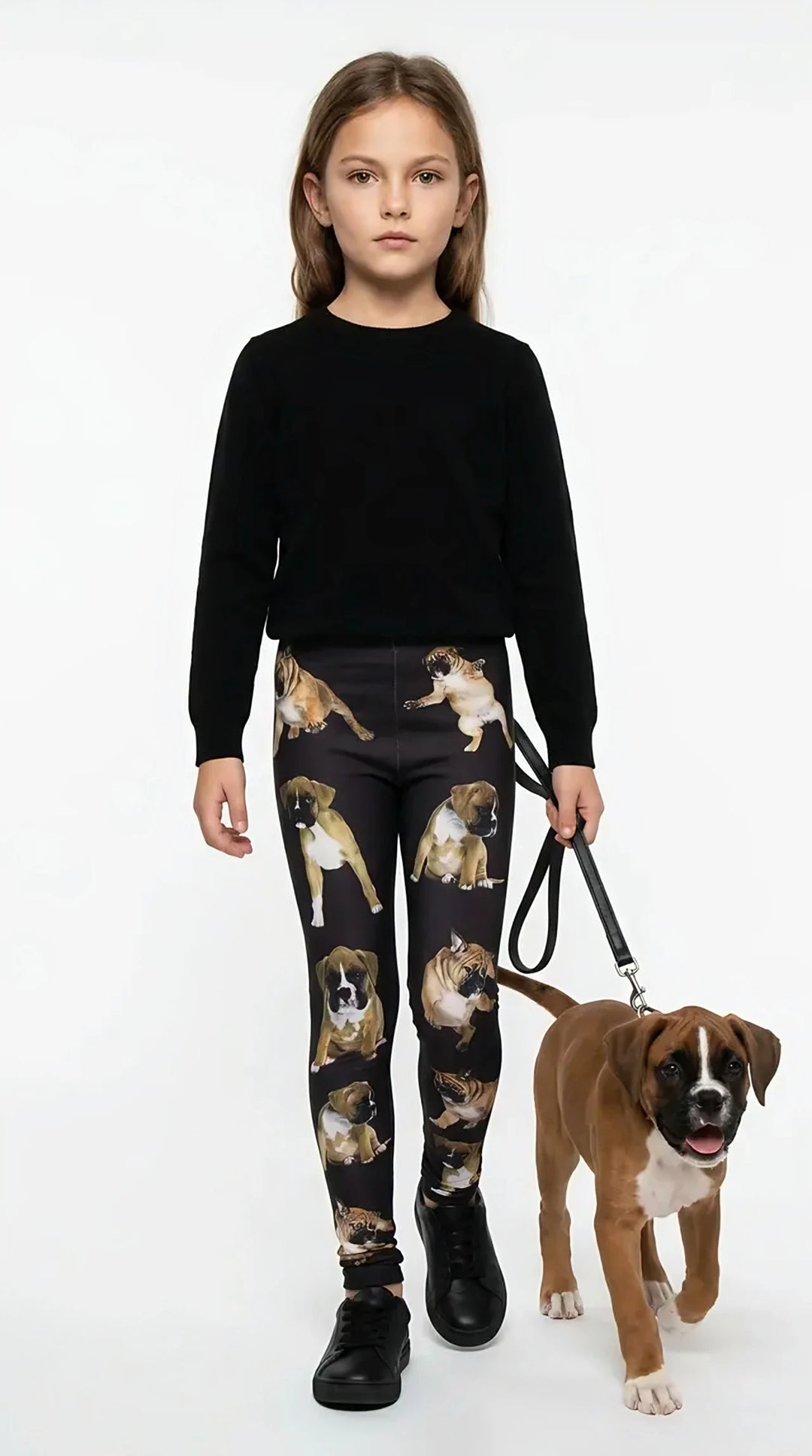 Girl in a black top and sneakers playing with a Boxer puppy outdoors, wearing black kids leggings with an all-over Boxer print