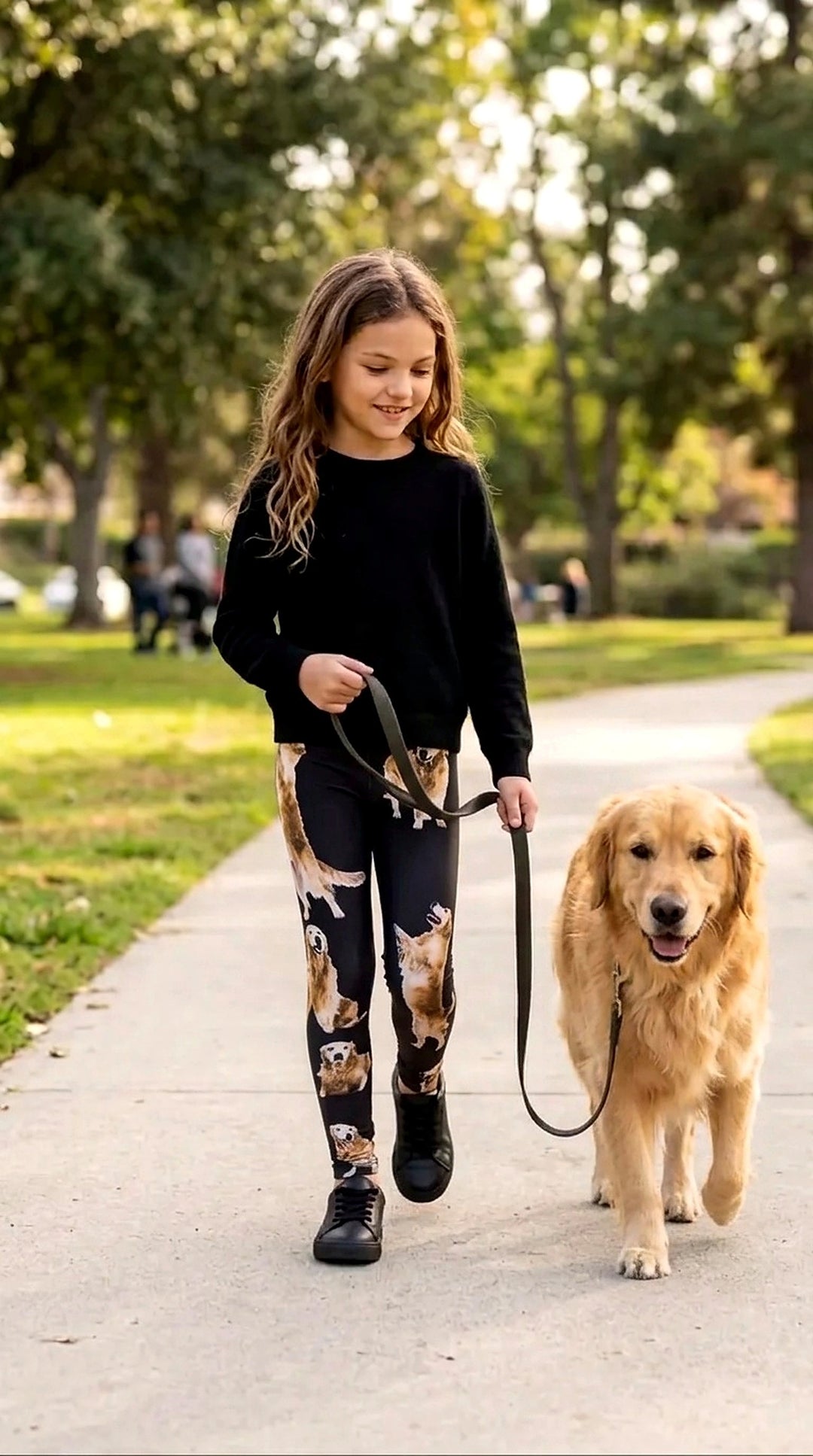 Smiling girl in a black sweater and black Golden Retriever print leggings walking a real Golden Retriever on a leash along a park path.