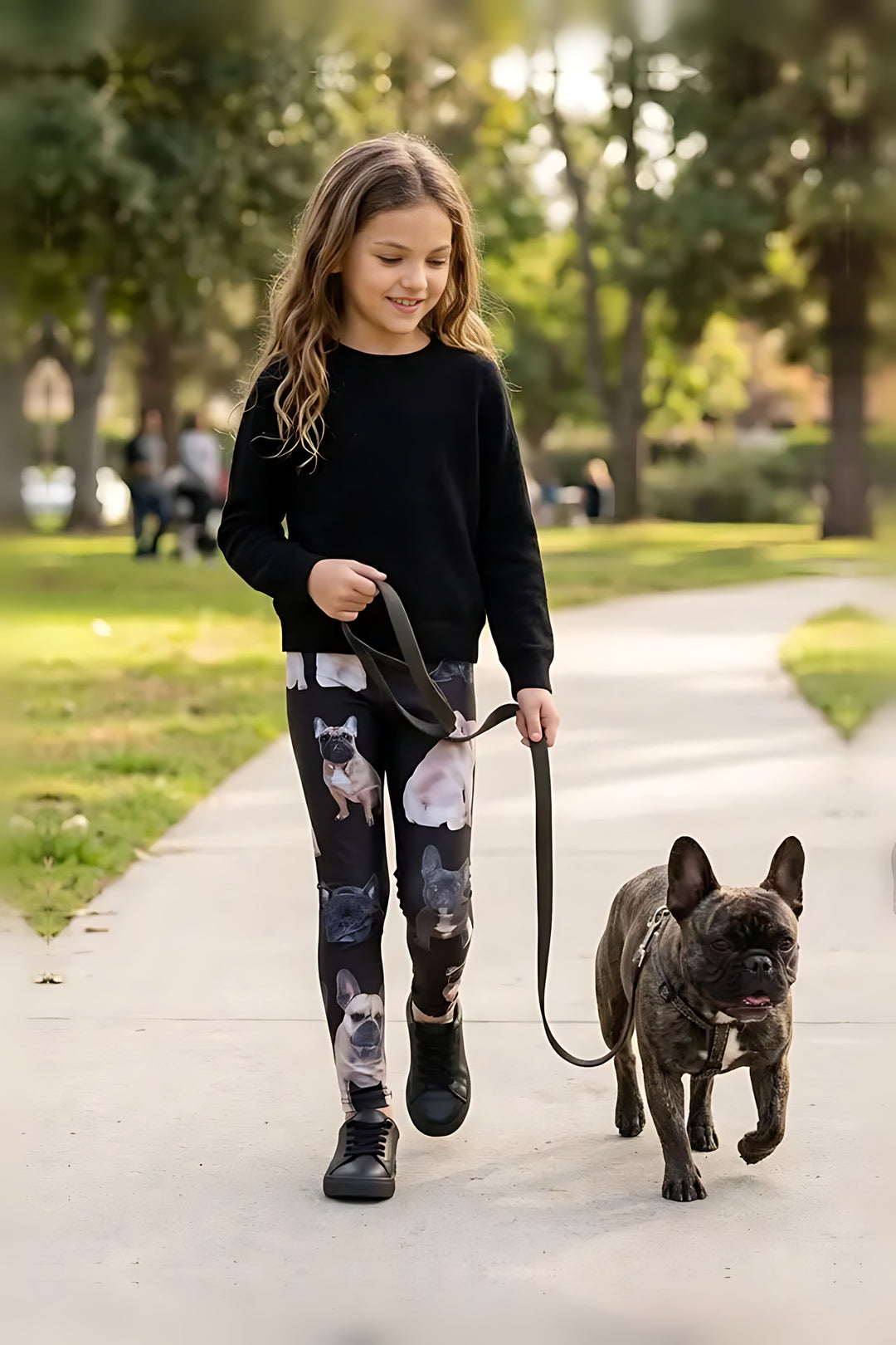 Girl in a black sweater walking a brindle French bulldog in a park, wearing French bulldog print girls leggings