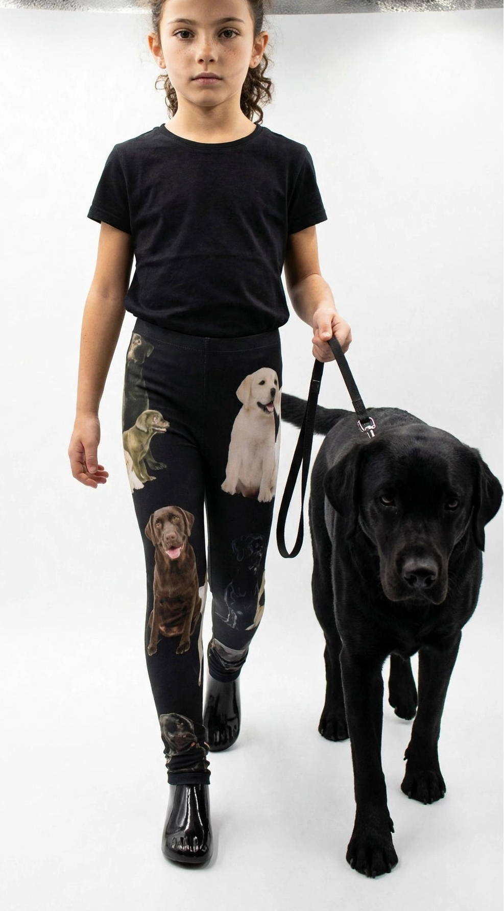 Young girl walking a real black Labrador on a leash while wearing Denimaxx kids Labrador print leggings and a black t-shirt in a white studio.