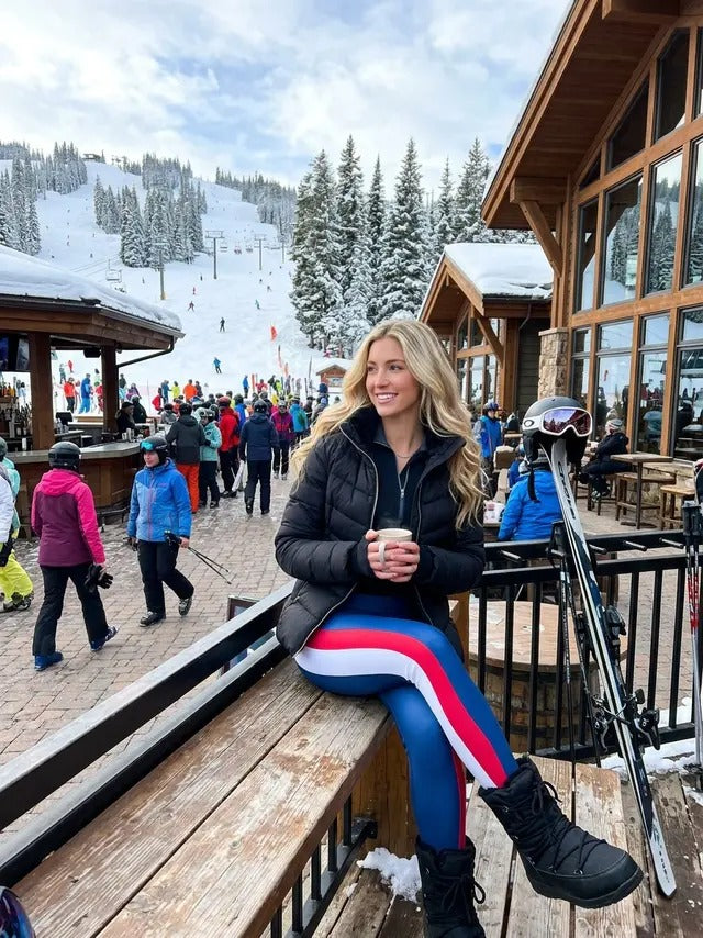 Woman sitting on a ski lodge deck with a mug, skis beside her, wearing Denimaxx royal blue leggings with a red-and-white racing stripe.
