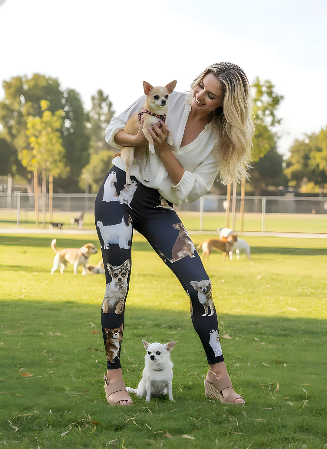 Woman holding a tan chihuahua and smiling, wearing black chihuahua print leggings, with a white chihuahua at her feet at a dog park.